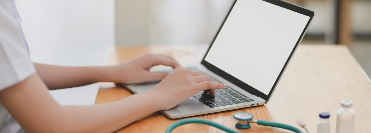 A person typing on a laptop with a stethoscope on the desk representing asking for a doctor's note when home sick from work