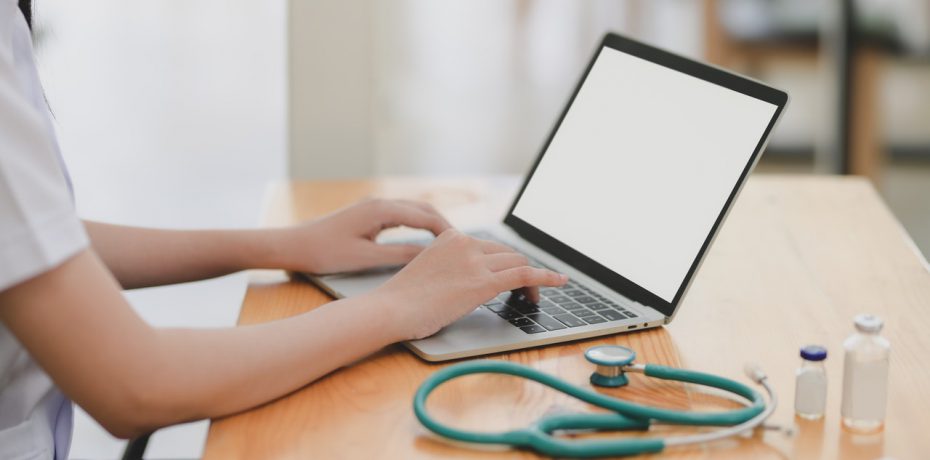 A person typing on a laptop with a stethoscope on the desk representing asking for a doctor's note when home sick from work