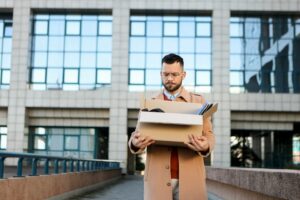 A man, terminated from his job, looks down with an expression of defeat as he carries a box of his belongings and walks away from the office building behind him, representing bonus and stock options after termination.