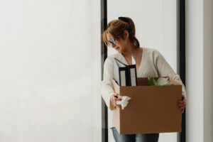 A woman, terminated from her job, carries a box of her belongings, with two binders and a plant poking out. She clutches a tissue in one hand, pinned between her finger and the box, and she is looking down and to the side with a sad frown, representing bonus and stock options after termination.