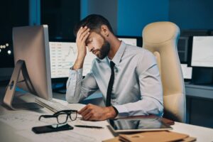 A man sits behind his desk in an office with his eyes closed and his forehead resting on his hand, expressing the workplace frustration that can lead to constructive dismissal.