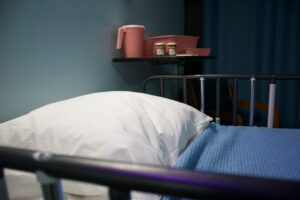 The top end of an empty hospital bed, with a pillow and blue blanket, and pill bottles and a plastic jug, and a basin on a table next to it, representing being fired after a sick leave in Ontario.