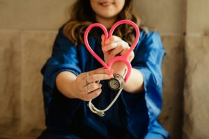 A healthcare worker wearing blue scrubs leans against a concrete wall while holding out a stethoscope so the tubes form a heart, representing workplace violence protection.